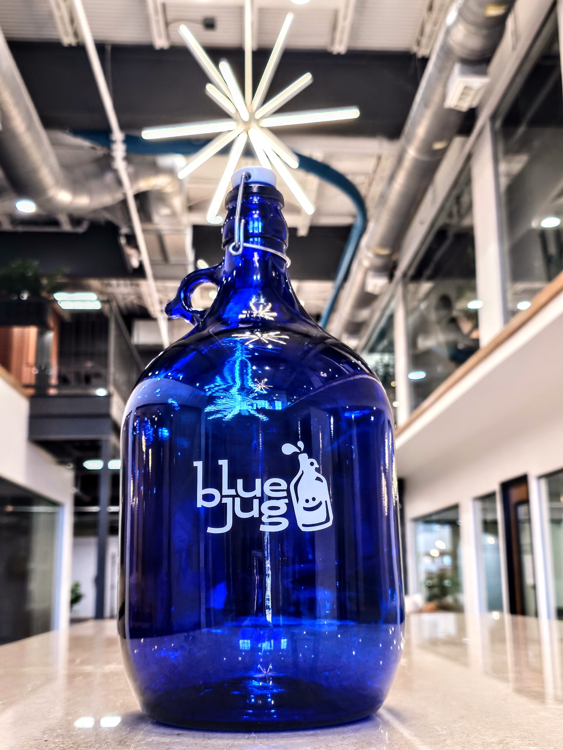 Close-up of a blue glass refillable water jug featuring the Blue Jug logo in a modern, brightly lit office or store setting.